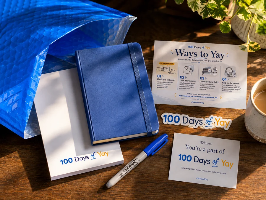 The Yay Kit unboxed on a wooden table: blue mailer, navy journal, 100 Days of Yay notepad, Ways to Yay guide card, Sharpie, sticker and welcome card.